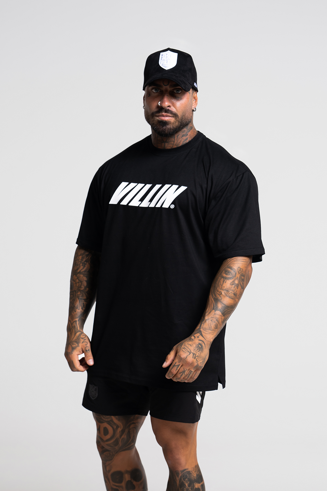 A man wearing a black oversized t-shirt with the brand name 'VILLIN' printed in large letters across the chest. The t-shirt is paired with black shorts and the person is also wearing a black cap.